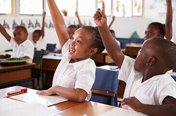 Young students in uniform, smiling and holding up their hands in a classroom - Teacher recruitment
